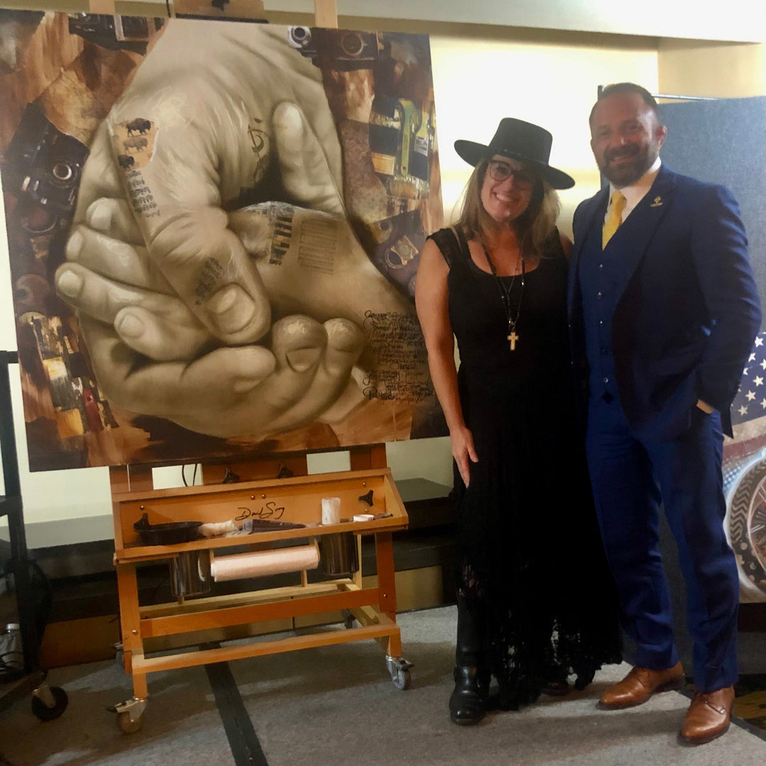 Two people posing in front of a large painting of hands on an easel.