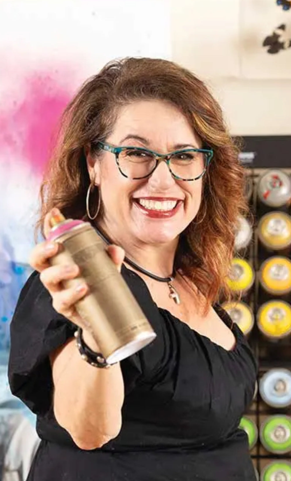 Artist ShenShen smiling spraying spray paint at the camera beautiful portrait wearing black in front of a white wall with a spray paint holding shelf
