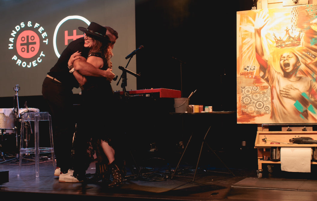 Two people, Tim Tebow and Shen embracing on a stage with a 'Hands & Feet Project' logo in the background next to the painting she did live