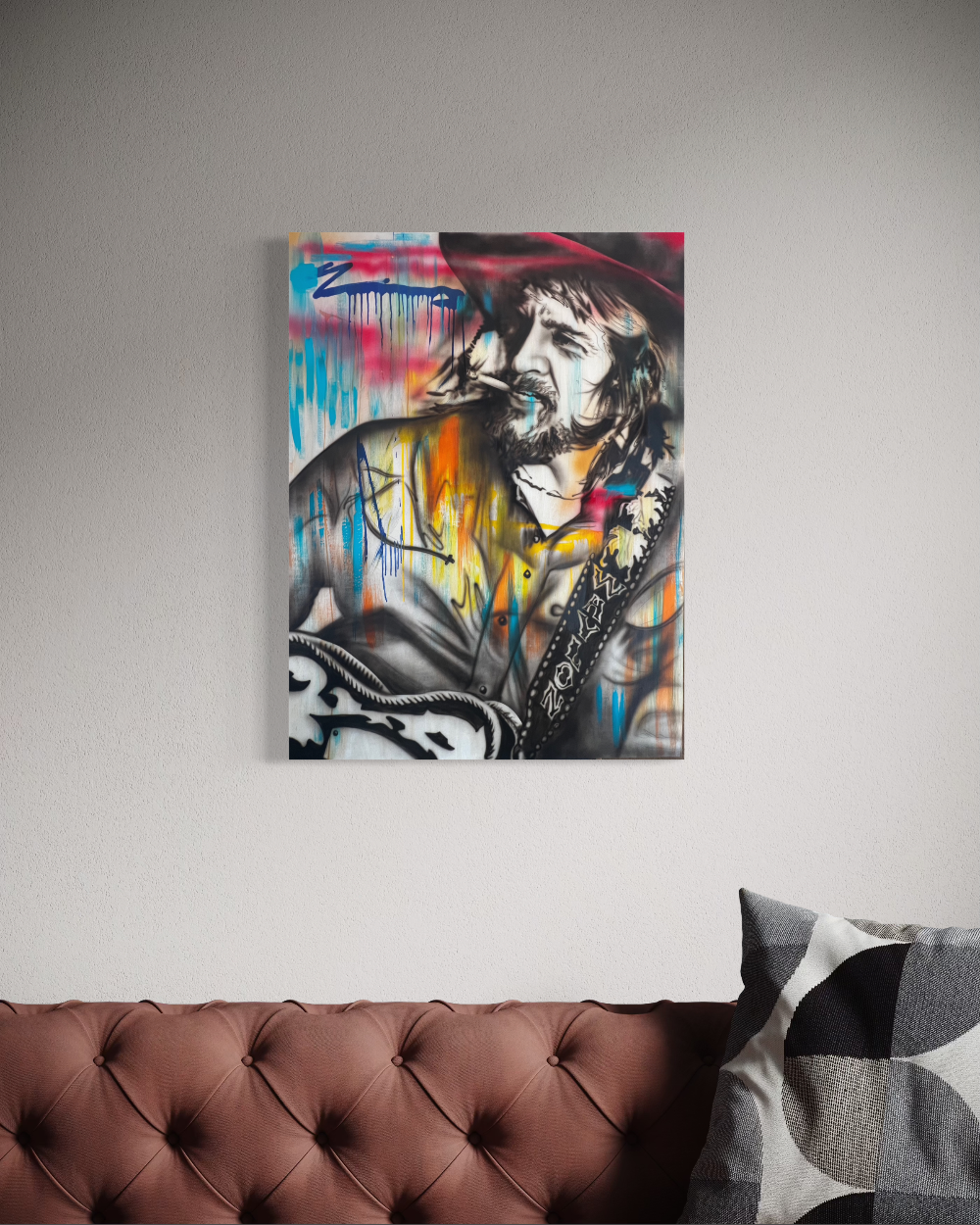 Colorful abstract painting of a person with a hat on a gray wall above a brown sofa. Contemporary portrait of Waylon Jennings playing guitar, featuring expressive black-and-white realism layered with vibrant splashes of blue, orange, pink, and yellow paint drips, celebrating the legendary outlaw country musician.